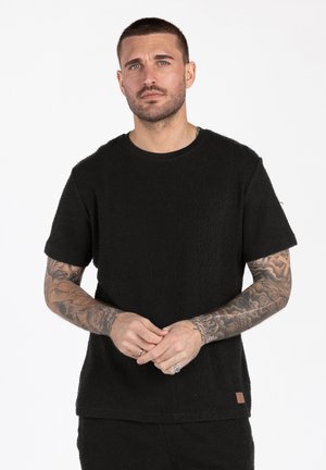 Mann mit kurzem Haar und Tätowierungen auf beiden Armen, der ein schwarzes, strukturiertes Kurzarmhemd trägt und die Hände vor sich verschränkt.