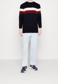 Maglione a coste blu navale con strisce orizzontali bianche e bordeaux, abbinato a jeans dritti azzurri chiari e sneaker scuri.