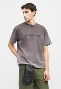 Νεαρός άνδρας που φοράει γκρι ξεβαμμένο μπλουζάκι Dickies και χακί παντελόνι, κρατώντας ένα μαύρο καπέλο το οποίο έχει τυλιγμένο στη ζώνη του.