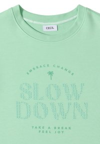 Sweat-shirt vert clair avec col côtelé, texte embossé « SLOW DOWN » et texte plus petit « Embrace Change », « Take a break, feel joy », plus un petit palmier.
