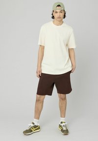 Chemise à manches courtes texturée crème, shorts marron et casquette verte. Pantalon de sport multicolore avec des accents jaunes et chaussettes blanches.