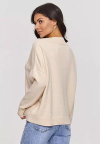 Beżowy, oversize sweter z ribbowanym dekoltem i dołem. Miękka tekstura, opadające ramiona i luźny krój. W zestawie z jasnoniebieskimi dżinsami.