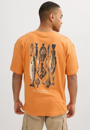 Hombre con camiseta naranja con múltiples ilustraciones detalladas de peces y texto "West Coast Anglers Society" en la parte trasera, pantalones cortos cargo beige.