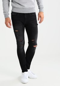 Pantalones vaqueros ajustados negros con múltiples áreas desgastadas. Combinados con zapatillas blancas y una sudadera gris, creando un conjunto casual.