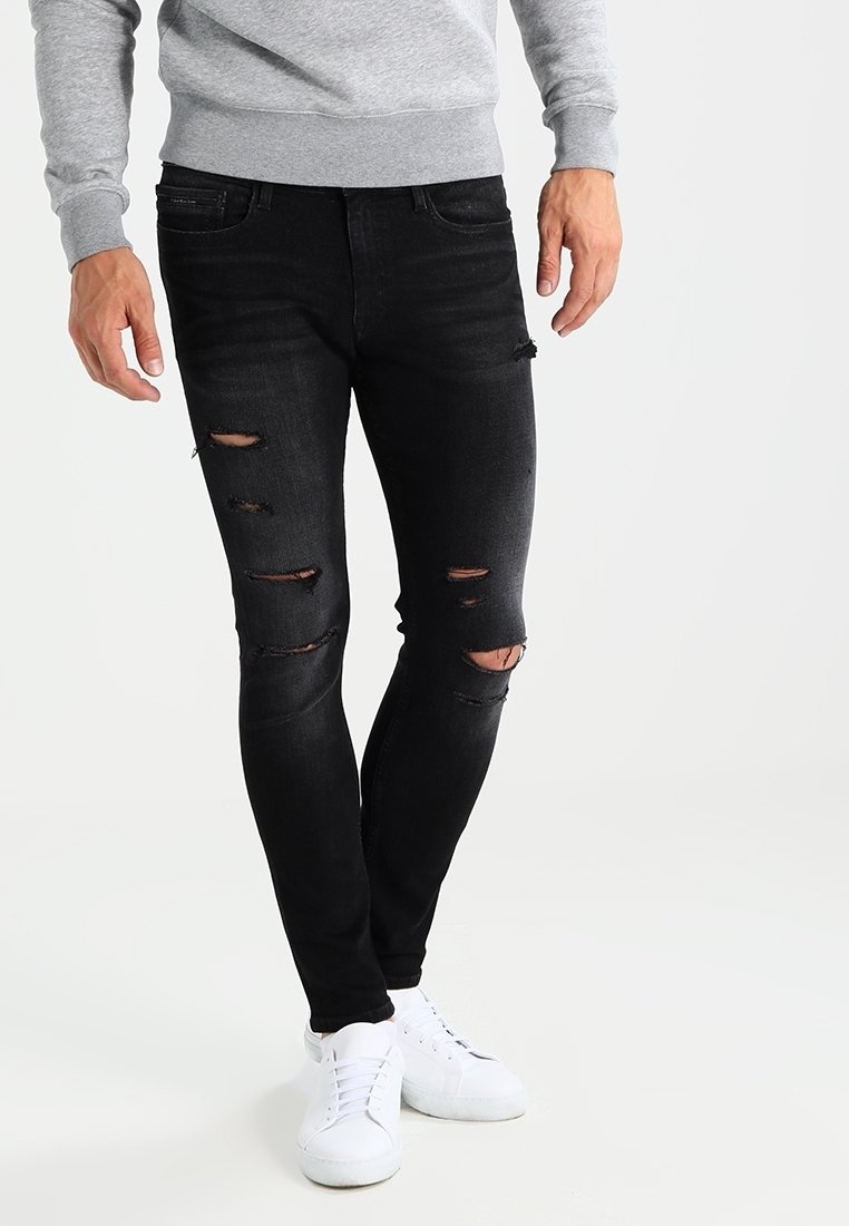 Pantalones vaqueros ajustados negros con múltiples áreas desgastadas. Combinados con zapatillas blancas y una sudadera gris, creando un conjunto casual.