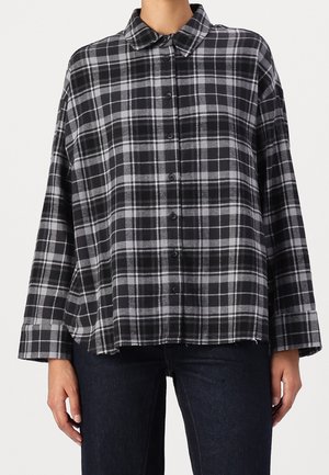 Vrouw draagt een losse zwart-grijs geruite blouse met kraag en knopen en donkere broek, staand tegen een witte achtergrond.