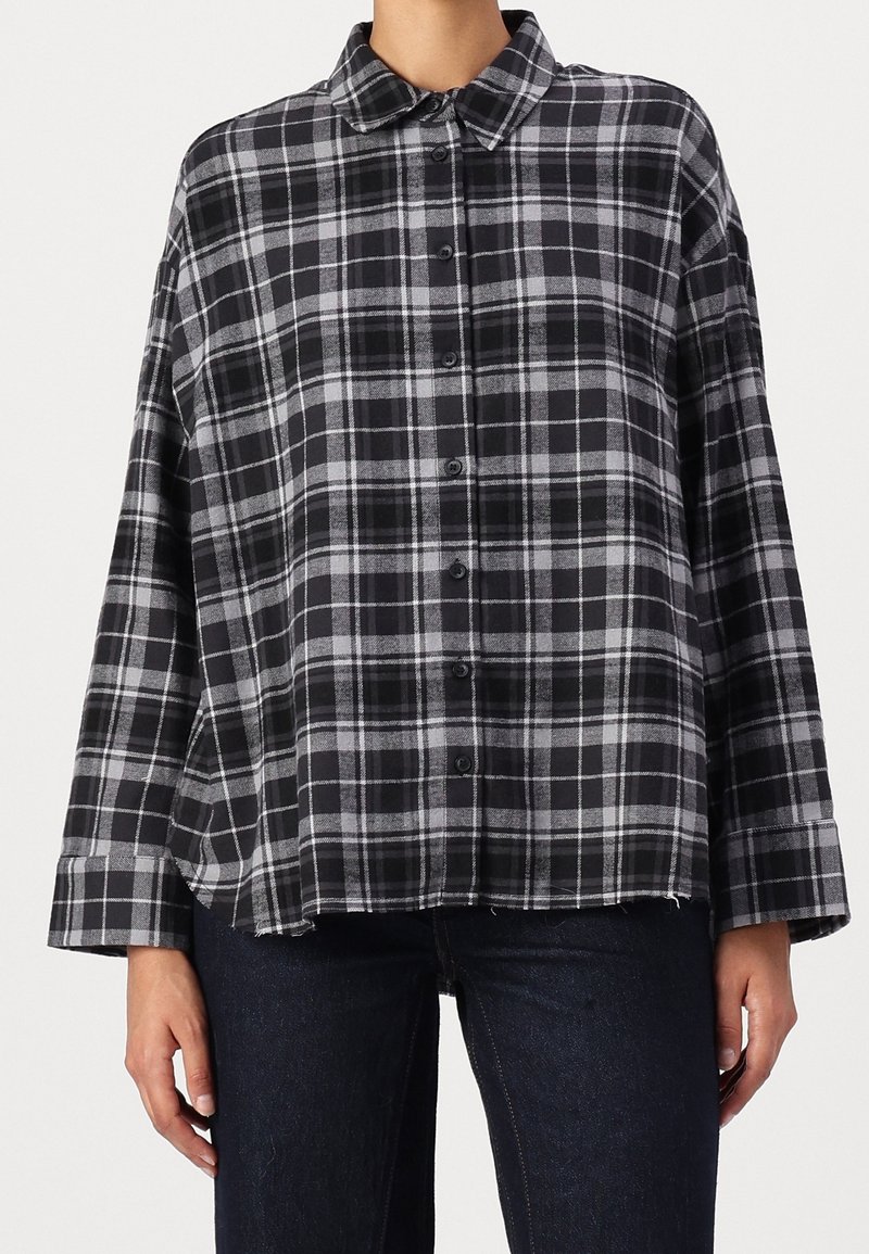 Femme portant une chemise ample à carreaux noirs et gris avec col et boutons, et un pantalon foncé, debout sur fond blanc.