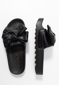 Black slip-on sandals with textured sole and large knotted bow on the upper strap, shown top and side views on white background.