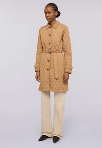 Gesteppter beiger Mantel mit tailliertem Gürtel, Knopfverschluss und langen Ärmeln. Kombiniert mit cremefarbenen Hosen und spitzen schwarzen Schuhen.