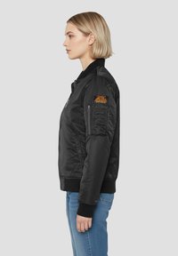 Czarna kurtka bomberka z suwakiem z przodu, ściągaczami na rękawach i kołnierzu. Na lewym rękawie znajduje się pomarańczowa naszywka. Gładki materiał i dopasowany krój.