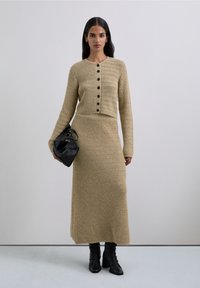 Woman wearing beige knit buttoned cardigan and matching long skirt, holding black leather handbag, and wearing black ankle boots.
