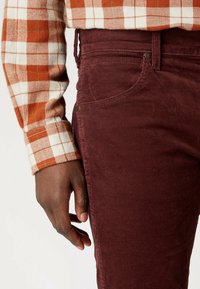 Vinröda kordfløyelbyxor med synlig ficksyddning och metallknapp. Skjorta av flanell med orange och vit rutig mönster.