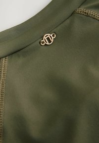 Detailný záber olivovo zelenej látky s detailným prešitím a malým okrúhlym zlatým kovovým logom pripevneným pri šve.