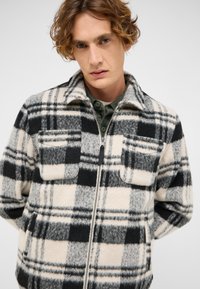 Kurtka z mieszanki wełny o czarno-białym wzorze w kratę, z zamkiem błyskawicznym oraz dwoma kieszeniami na piersi z zaokrąglonymi klapkami.