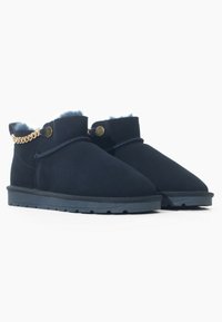 Botines de ante azul marino con un suave forro de piel azul, un acento decorativo de cadena dorada y una suela de goma negra texturizada.