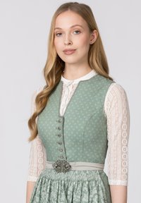 Hellgrünes Dirndl mit Blumenmuster, silbernen Knöpfen und geflochtenem Taillengürtel. Mit einer weißen Spitzenbluse mit Dreiviertelärmeln.