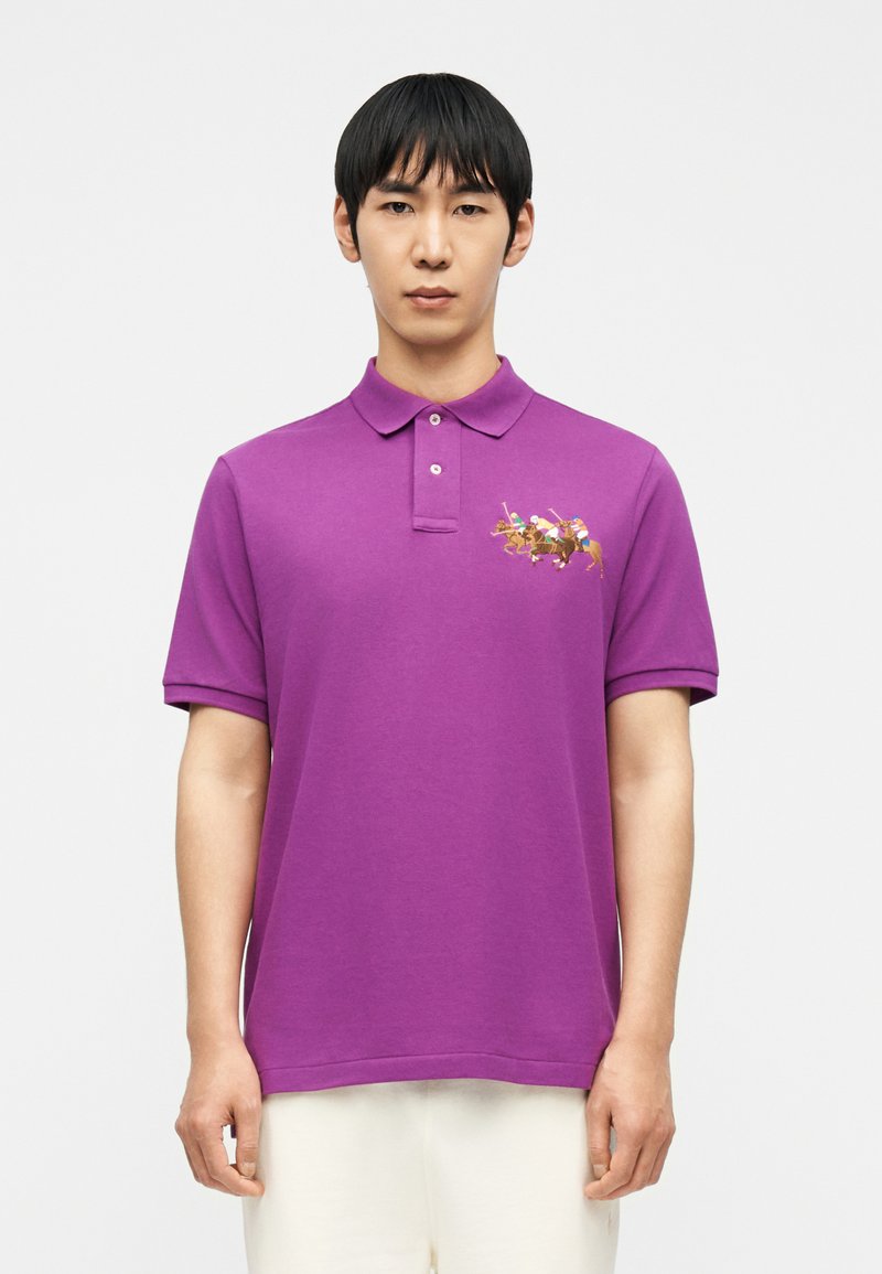 Άνδρας που φοράει μοβ πόλο με πολύχρωμο κέντημα  παίκτη πόλο στο αριστερό στήθος, συνδυασμένο με λευκό παντελόνι.