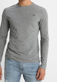 T-shirt grigio a maniche lunghe realizzato in tessuto morbido, con collo tondo e un piccolo logo sul petto sinistro. Abbinato a jeans azzurri chiari.