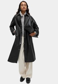 Černý kožený kabát s patentkami, oversize střih a široké rukávy. V kombinaci s šedým svetr s límcem a světlými béžovými kalhotami s širokými nohavicemi.
