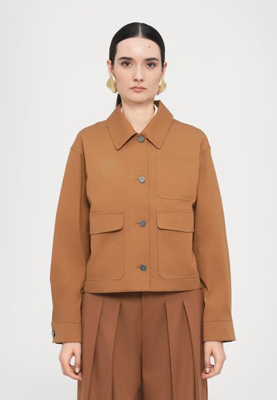 Veste marron courte avec un col évasé, fermeture à boutons sur le devant, deux grandes poches et boutons gris. Portée sur un pantalon plissé marron assorti.
