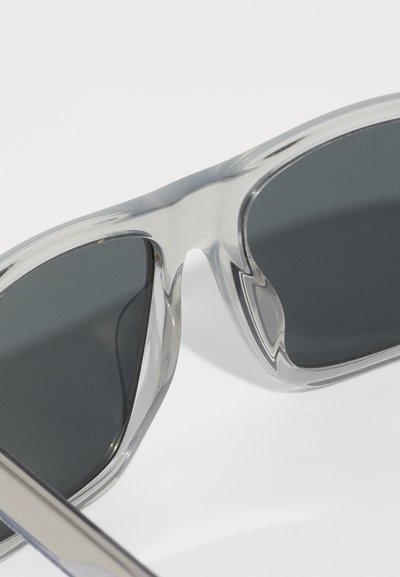Ochelari de soare din plastic transparent cu lentile gri închis. Designul unghiular al ramei are margini netede și un finisaj lucios, sporind durabilitatea.