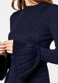 Marineblauer Pullover mit langen Ärmeln, hohem Kragen und ausgestellten, gefältelten Manschetten. Der Pullover zeichnet sich durch einen strukturierten, glitzernden Stoff aus.