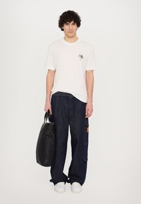 Camiseta blanca de manga corta con estampado gráfico, combinada con pantalones de denim holgados de color azul oscuro que cuentan con un bolsillo lateral y una etiqueta marrón. Bolsa de mano negra.