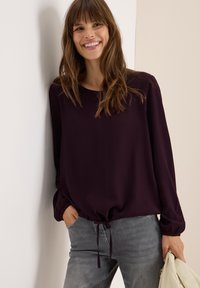 Blouse violet foncé à manches longues, col rond et d�étail à nouer à l'ourlet, associée à un jean gris clair. Tissu lisse avec des rayures verticales.