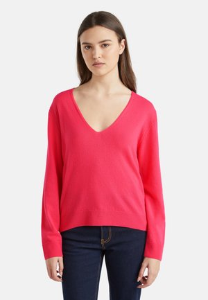 Suéter de pico de color rosa brillante, con mangas largas, dobladillo acanalado y una textura suave. Combinado con jeans azul oscuro.