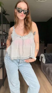 Top cinza claro com rendas e detalhe em botão, combinado com jeans de denim azul claro, em um ambiente interior iluminado.