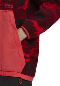 Röd och svart mönstrad fleecepullover med en slät röd ficka, elastiska ärmmuddar och dragsko i nederkant. Inkluderar logobroderi på bröstet.