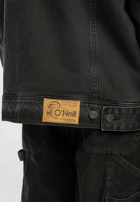 Svart denimjacka med beige O'Neill-märke och metallknappar, kombinerat med svarta texturerade byxor med sidfickor.