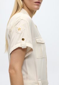 Femme blonde portant une chemise beige à manches courtes avec un écusson étoile dorée et un détail de bouton sur la manche ainsi qu'une poche poitrine à l'avant.