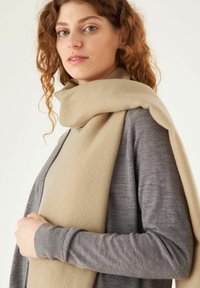 Écharpe beige drapée sur un cardigan gris. L'écharpe a une texture douce ; le cardigan présente un design simple et ajusté avec des manches longues.