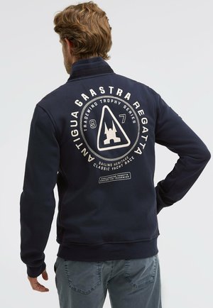 Mann mit hellbraunem Haar, der eine marineblaue Jacke mit weißem, segelthemenbezogenem Text und Logo auf dem Rücken trägt und vor einem schlichten Hintergrund steht.