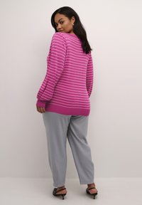 Magenta Pullover mit weißen Streifen, mit puffigen Ärmeln und einem gerippten Saum. Kombiniert mit hellgrauen Hosen und schwarzen High Heels.
