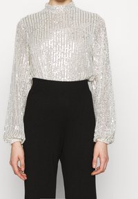 Blusa leggera e tinta unita con paillettes e righe verticali argento, colletto alto e maniche a sbuffo; abbinata a pantaloni neri a vita alta.