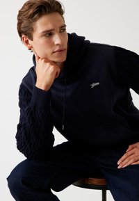 Felpa blu marina con maniche a treccia testurizzate, cappuccio con cordino e piccolo logo ricamato sul petto, indossata con jeans in denim scuro.