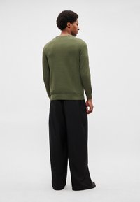 Alyvuogių žalios spalvos megztinis su apvalia kaklo linija, tekstūruotu paviršiumi ir apsiūtais rankogaliais, derinamas su plačiomis juodomis kelnėmis, matomas iš nugaros.