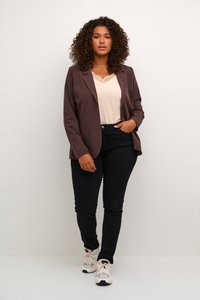 Blazer marron à bouton unique, porté sur un haut crème avec des détails en dentelle. Assorti avec un jean noir et des chaussures de sport blanches.