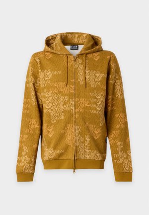 Jeune homme portant un hoodie zippé jaune moutarde avec un texte imprimé, jean large bleu clair, baskets blanches, debout devant un fond uni.