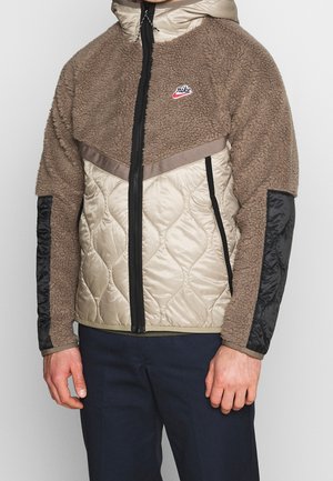 Homme portant une veste Nike marron et beige avec un devant matelassé, un corps supérieur et des manches duveteux, des coudières noires et une fermeture éclair à l'avant.