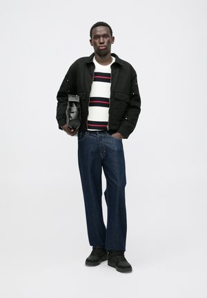Veste noire, pull rayé blanc et rouge, jeans larges bleus et chaussures noires. Tient un petit sac noir. Design simple et moderne.