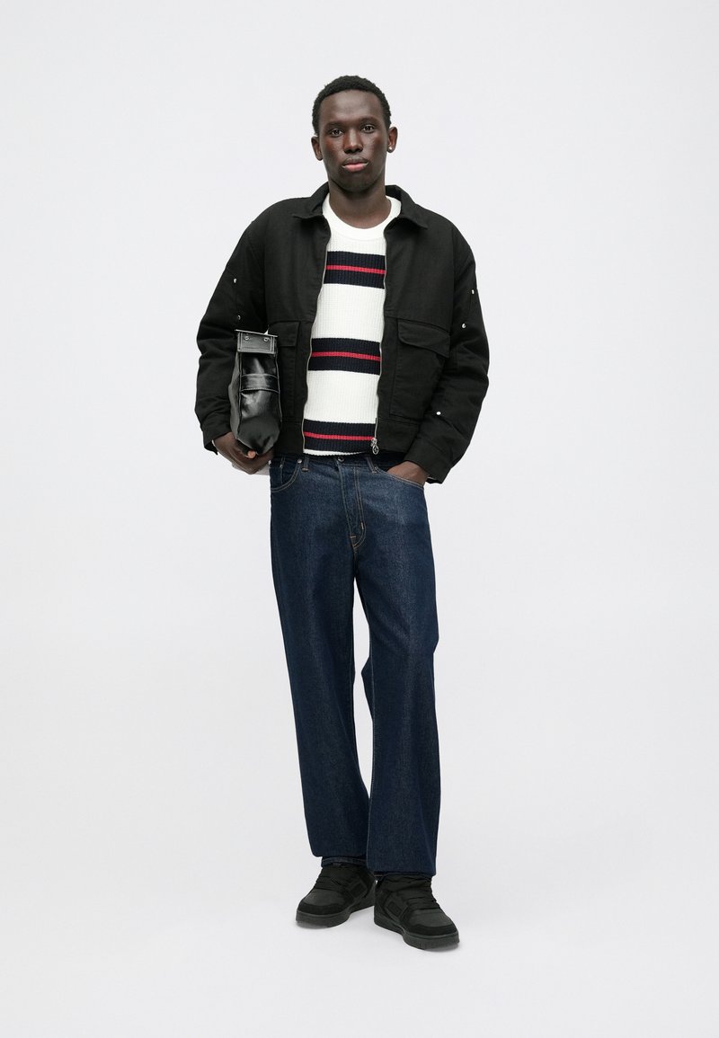 Chaqueta negra, suéter a rayas blancas y rojas, jeans de pierna ancha azules y zapatos negros. Sosteniendo un pequeño bolso negro. Diseño simple y moderno.