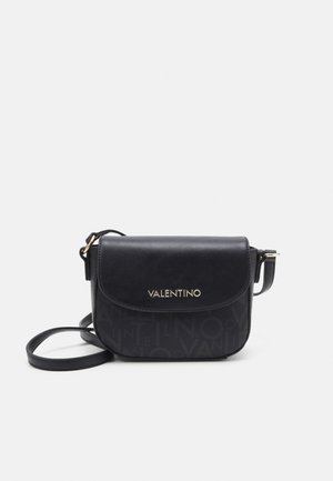 Černá crossbody taška s hladkým povrchem a zlatým logem. Má překlopné zavírání a dlouhý popruh pro nošení přes tělo.