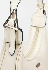 Närbild av en krämfärgad handväska med texturerat band, guldtriangulär spänne och svart kantdetalj på en slät yta.