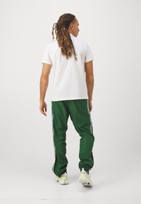 Maglietta bianca a maniche corte abbinata a pantaloni da pista verdi con strisce bianche ai lati e polsini con zip, completata da sneakers bianche.