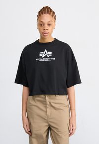 Juodas trumpas maikė, pagaminta iš medvilnės, su balta Alpha Industries logotipu ir geometriniais akcentais priekyje. Papildyta chaki spalvos kelnėmis.