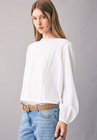 Blusa bianca, con texture, maniche lunghe a sbuffo e dettagli in pizzo lungo il colletto e il davanti; abbinata a jeans azzurri e una cintura marrone.