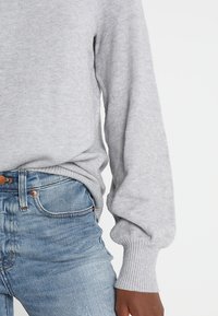 Hellgrauer Pullover mit gerippten Bündchen und Saum, in lockerer Passform. Über blauen Jeans mit einem Kupferknopf-Detail getragen. Weiche Textur.